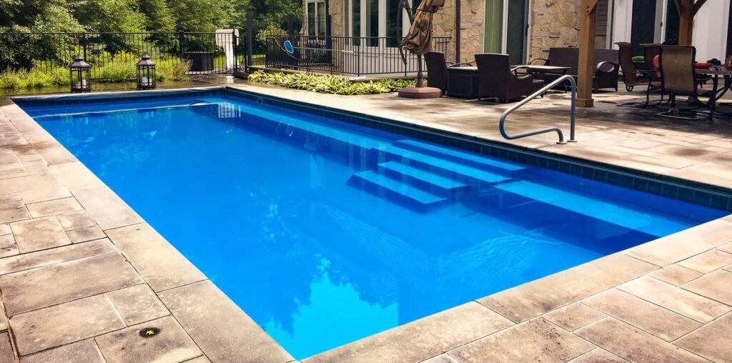 A backyard with a rectangular fiberglass pool surrounded by stone pavers, outdoor seating, and greenery.
